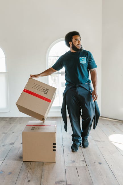 A man with dark curly hair and a beard, dressed in a blue T-shirt and dark trousers, is inside a bright, empty room with light-colored walls and wooden flooring. He is holding a cardboard box with red tape around it, preparing to place it on top of another box on the floor. The background features a large arched window allowing natural light to fill the space. This scene depicts the packing process during home relocation, characteristic of furniture transport and moving logistics services provided by Edgware Man and Van. The image captures the careful handling of packaging materials, essential in safe moving and packing operations during house removals, illustrating the practical aspects of loading and organizing items within a residential property in preparation for transportation.