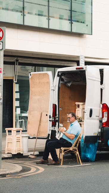A man wearing glasses and a light blue shirt is seated on a wooden chair beside an open white moving van parked on a city street. Inside the van, visible are large cardboard boxes and wooden furniture pieces, likely packed for a house relocation. The van's rear doors are open, revealing additional packed items, with the interior illuminated by natural daylight. A wooden panel leans against the van’s side, and nearby on the pavement, there are plastic wrapping and protective materials prepared for furniture transport. In the background, a modern building with glass windows and metal panels, along with a street sign, indicates an urban environment. The scene captures the loading or unloading process typical of professional removals—such as those offered by Edgware Man and Van—highlighting the logistical aspects of packing, furniture transport, and home relocation activities.