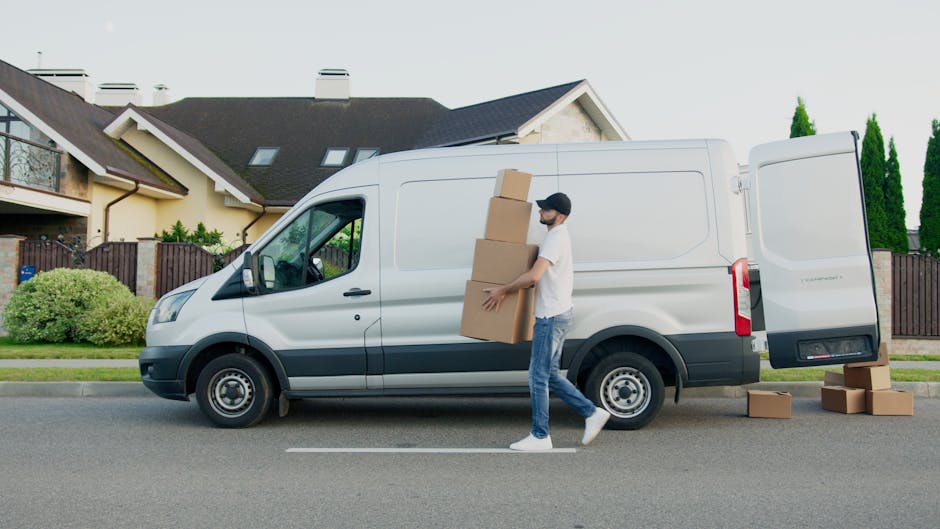 A man dressed in casual clothes, wearing a black face mask, is carrying three cardboard boxes stacked vertically outside on a residential street during daytime. He is walking beside a white commercial van parked parallel to the curb, which is used for home relocation and furniture transport, with the van's rear door open. The van is positioned in front of a modern two-storey house with a dark roof, light-colored walls, and large windows, surrounded by a wooden fence and landscaped greenery including bushes and trees. Several additional cardboard boxes are placed on the pavement near the rear of the van, ready for loading or unloading. The scene depicts a process of packing and moving, highlighting the logistics involved in house removals, with the van serving as the main transport vehicle for furniture and packing materials in a residential environment.