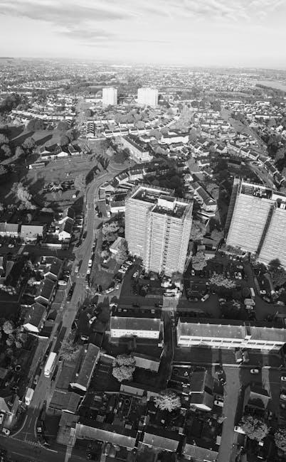 A black and white aerial photograph of a residential area featuring a mix of multiple high-rise apartment buildings and smaller houses. The high-rise buildings are situated near the centre of the image, with parking areas and roads surrounding them. In the foreground, narrow streets are lined with terraced houses, some with small gardens and trees visible from above. The streets are populated with parked cars and moving vehicles, and the overall scene includes a combination of urban housing and infrastructure. The background shows a vast expanse of cityscape stretching toward the horizon under a slightly cloudy sky. This image illustrates an urban environment suitable for home relocation or furniture transport services, as seen from the perspective of a professional removals company's deployment area. Edgware Man and Van operates in locations like Hale Lane flats, as indicated on the webpage for house removals and moving logistics.