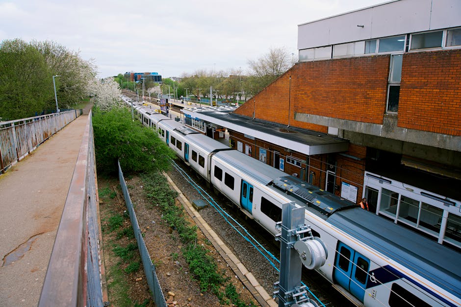 A view from an elevated pedestrian walkway overlooking Edgware station with a train on the tracks below. The train has white and blue carriages and is positioned parallel to the platform, which is partially visible beneath a brick and metal station building. The station building features a flat roof and large windows; some windows are open. The platform edge has safety barriers, and there are various signs and notices along its length. To the left, there is a metal railing separating the walkway from the railway tracks, with some greenery and small flowering trees growing alongside. The surrounding environment includes a green landscape with trees and overcast sky, typical of urban railway stations. This setting showcases a typical scene where home relocation services by Edgware Man and Van might coordinate furniture transport and packing efforts during a moving process involving station access considerations, highlighting the loading and unloading areas close to the train station.