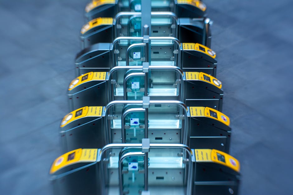 A row of eight luggage trolleys with yellow barcode scanners attached to their handles, positioned on a grey tiled floor indoors. The trolleys are made of metal with four wheels each, and some have transparent plastic bags draped over their frames. The lighting is bright and even, highlighting the reflective surfaces of the trolleys. This setup appears to be in a logistics or transportation environment, possibly within a station or warehouse, supporting the process of home relocation or furniture transport. The image aligns with the context of packing and moving services provided by Edgware Man and Van, as indicated on the webpage about Edgware station moving access times for removals.