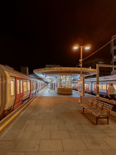 A view from an elevated pedestrian walkway overlooking Edgware station with a train on the tracks below. The train has white and blue carriages and is positioned parallel to the platform, which is partially visible beneath a brick and metal station building. The station building features a flat roof and large windows; some windows are open. The platform edge has safety barriers, and there are various signs and notices along its length. To the left, there is a metal railing separating the walkway from the railway tracks, with some greenery and small flowering trees growing alongside. The surrounding environment includes a green landscape with trees and overcast sky, typical of urban railway stations. This setting showcases a typical scene where home relocation services by Edgware Man and Van might coordinate furniture transport and packing efforts during a moving process involving station access considerations, highlighting the loading and unloading areas close to the train station.