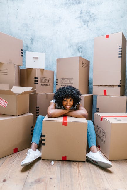 A young woman with curly black hair, dressed casually in a white top, blue jeans, and white sneakers, is sitting on the floor inside a room surrounded by numerous cardboard moving boxes. She is smiling happily and leaning forward with her arms resting on a large box, which is sealed with red packing tape. The boxes are stacked against the pale blue, slightly textured wall and are of varying sizes, some partially open, revealing packing materials such as tissue paper and bubble wrap. The scene is well-lit with natural light, highlighting the wooden floor and the clutter of packing supplies. This image depicts a home relocation or packing process, associated with furniture transport and moving logistics handled by Edgware Man and Van during a same-day removal service to avoid delays and extra fees, as referenced on the webpage about removals in Edgware.