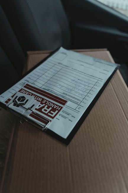 A shipping and handling form placed on top of a cardboard box inside a vehicle, with part of a darker seat visible in the background. The form is secured with a clip featuring a warning label that reads 'FRAGILE HANDLE WITH CARE' in bold red letters. The cardboard box underneath is unlabelled and appears to be part of a home relocation; it is positioned on the vehicle's interior floor, adjacent to other moving materials. The lighting is natural and soft, suggesting the vehicle is stationary. This scene depicts the packing and loading process involved in house removals, as provided by Edgware Man and Van, highlighting the careful handling of items during furniture transport and home move logistics.