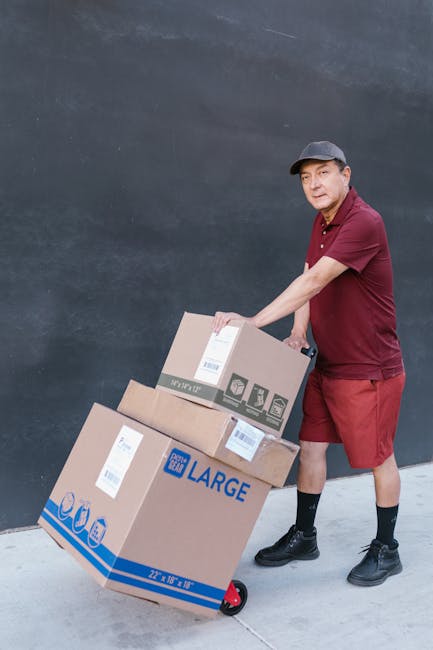 A young woman with curly black hair, dressed casually in a white top, blue jeans, and white sneakers, is sitting on the floor inside a room surrounded by numerous cardboard moving boxes. She is smiling happily and leaning forward with her arms resting on a large box, which is sealed with red packing tape. The boxes are stacked against the pale blue, slightly textured wall and are of varying sizes, some partially open, revealing packing materials such as tissue paper and bubble wrap. The scene is well-lit with natural light, highlighting the wooden floor and the clutter of packing supplies. This image depicts a home relocation or packing process, associated with furniture transport and moving logistics handled by Edgware Man and Van during a same-day removal service to avoid delays and extra fees, as referenced on the webpage about removals in Edgware.